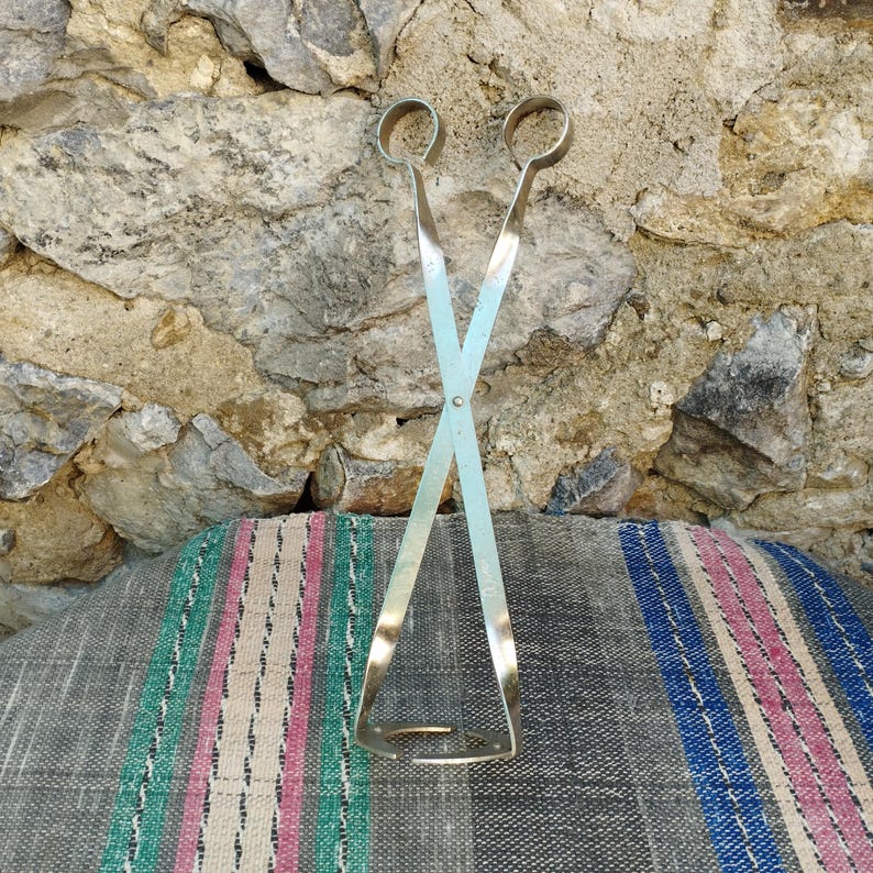May include: A pair of silver-toned metal tongs with a crossed design. The tongs are on a striped fabric background.