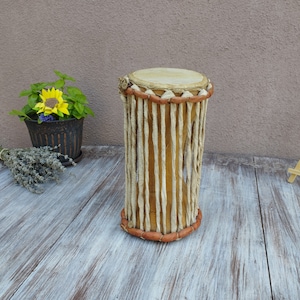 May include: A handcrafted djembe drum with a natural wood finish. The drum features a light-coloured drumhead, a woven body made of vertical strips, and decorative brown trim around the top and bottom. The drum is cylindrical and stands on a wooden surface.