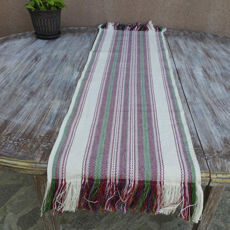 Pode incluir: Um caminho de mesa creme com listras verticais vermelhas, verdes e roxas. O caminho de mesa tem bordas com franjas de cores correspondentes. O tecido &eacute; exibido em uma mesa de madeira desgastada, com uma planta em vaso ao fundo.