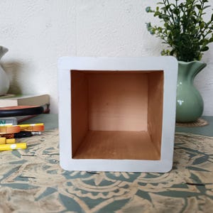 Puede incluir: Caja de madera blanca con un interior marr&oacute;n claro. La caja est&aacute; vac&iacute;a y tiene forma cuadrada.