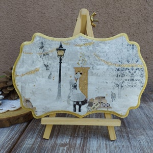 May include: A white and gold decorative plaque with a winter scene featuring a girl in a white dress and a black coat standing in front of a golden door. The plaque is on a small wooden easel.