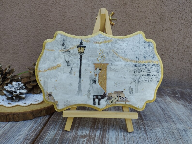 May include: A white and gold decorative plaque with a winter scene featuring a girl in a white dress and a black coat standing in front of a golden door. The plaque is on a small wooden easel.