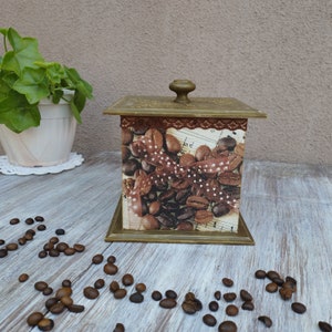 May include: A square wooden box with a brown lid and a gold trim. The box is decorated with a design of coffee beans and a musical note. The box is sitting on a wooden surface.