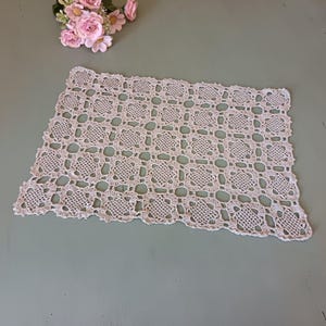 May include: A rectangular light pink crochet doily featuring a repeating square motif pattern with an openwork design and a scalloped edge. The textile is crafted with fine thread, creating a delicate grid of interconnected floral-like shapes.