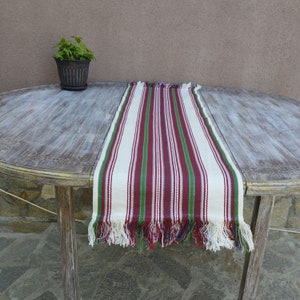 Pode incluir: Um caminho de mesa listrado em vermelho, verde e branco, com bordas com franjas. O caminho de mesa &eacute; exibido em uma mesa de madeira desgastada. Um pequeno vaso de plantas &eacute; vis&iacute;vel ao fundo.