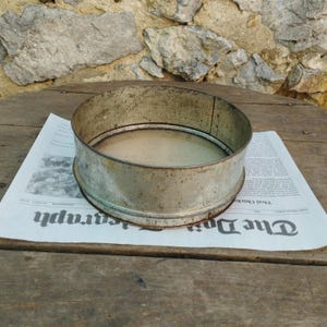 Op de afbeelding: Een vintage metalen zeef met een cirkelvormig ontwerp. De zeef is gemaakt van een dof, zilverkleurig metaal en heeft een fijnmazig scherm. Het ligt op een krant met zichtbare tekst. De zeef wordt waarschijnlijk gebruikt voor het zeven van bloem of andere droge ingrediënten.
