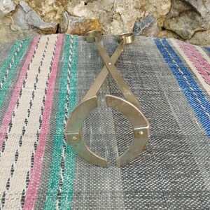 May include: A pair of silver metal bottle stoppers with a unique design. The stoppers are crossed and have a circular shape with a small loop at the top.
