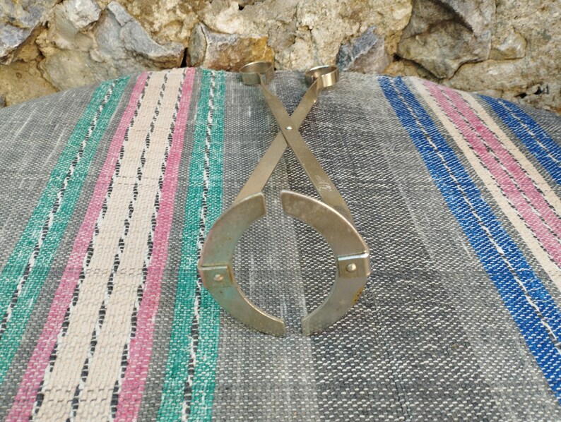 May include: A pair of silver metal bottle stoppers with a unique design. The stoppers are crossed and have a circular shape with a small loop at the top.