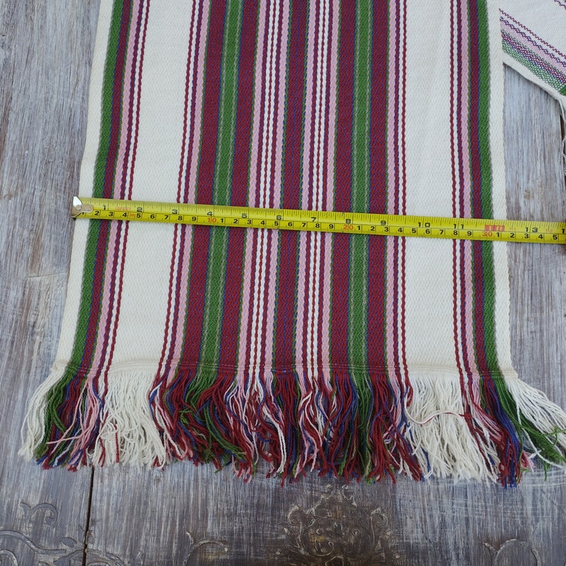 Pode incluir: T&ecirc;xtil creme com listras verticais vermelhas, verdes e rosa, com franjas na parte inferior. O t&ecirc;xtil tem aproximadamente 33 cm de comprimento. Uma fita m&eacute;trica amarela est&aacute; colocada sobre o t&ecirc;xtil.