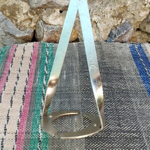 May include: A silver metal bottle holder with a circular base and two curved arms that meet at a point. The holder has the word "30 cm" stamped on one of the arms.