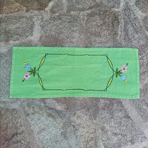 May include: A rectangular, light green hand towel with an embroidered floral design. The embroidery features pink and blue flowers with yellow accents, and a black outline. The towel is made of a soft, absorbent material.