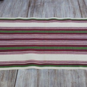 Pode incluir: Caminho de mesa creme com listras horizontais em tons de verde, bord&ocirc; e rosa. As listras s&atilde;o espa&ccedil;adas uniformemente e criam uma apar&ecirc;ncia texturizada. O caminho de mesa &eacute; feito de tecido e &eacute; exibido em uma superf&iacute;cie de madeira clara.
