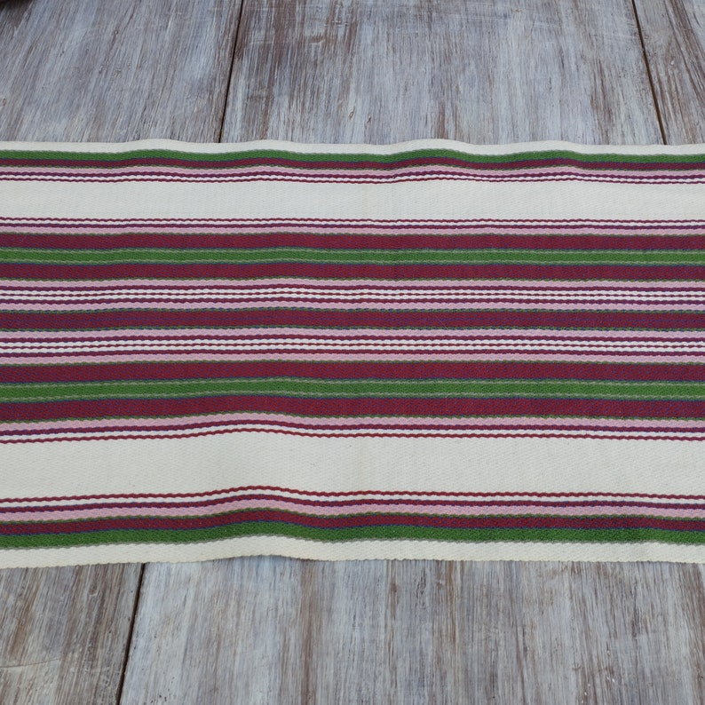 Pode incluir: Caminho de mesa creme com listras horizontais em tons de verde, bord&ocirc; e rosa. As listras s&atilde;o espa&ccedil;adas uniformemente e criam uma apar&ecirc;ncia texturizada. O caminho de mesa &eacute; feito de tecido e &eacute; exibido em uma superf&iacute;cie de madeira clara.