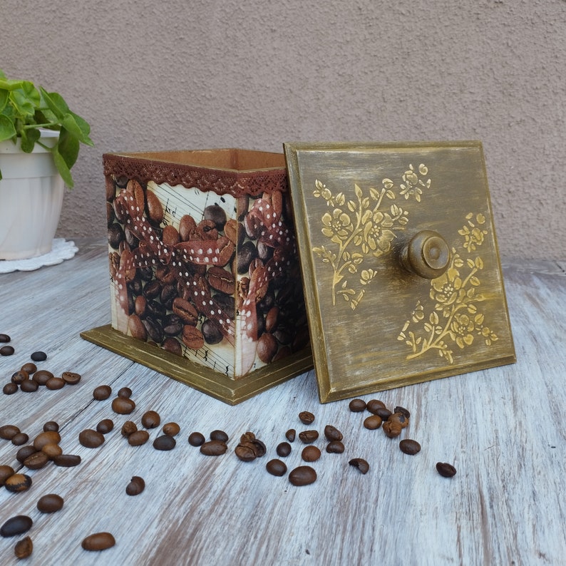 May include: A decorative wooden box with a floral design on the lid and a coffee bean pattern on the sides. The box is closed and sitting on a wooden surface.
