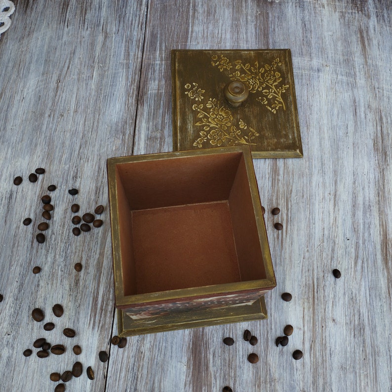 May include: A wooden box with a floral design on the lid. The box is open and the inside is empty. The box is sitting on a light wood surface.