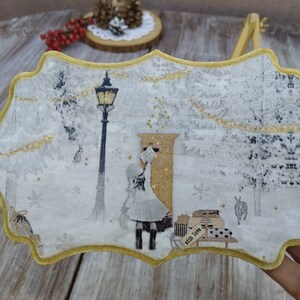 May include: A white wooden ornament with a gold trim, featuring a winter scene with a girl standing in front of a mailbox. The scene includes a street lamp, snow-covered trees, and the text "With Love & Joy".