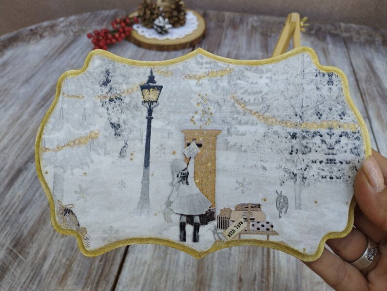 May include: A white wooden ornament with a gold trim, featuring a winter scene with a girl standing in front of a mailbox. The scene includes a street lamp, snow-covered trees, and the text "With Love & Joy".