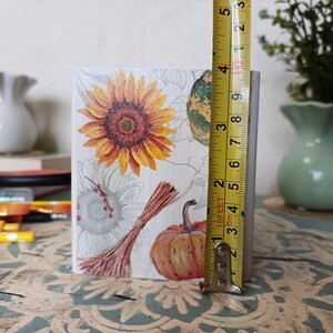 Puede incluir: Una caja de madera blanca con un dise&ntilde;o de oto&ntilde;o. La caja presenta un girasol, una calabaza y una calabaza. La caja mide 9 cm de alto.
