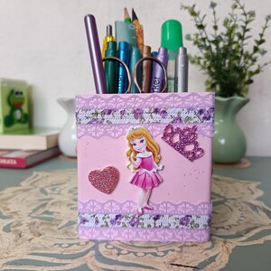 May include: Pink princess-themed pencil holder with a princess character, a crown, and a heart. The holder is decorated with lace and floral patterns.