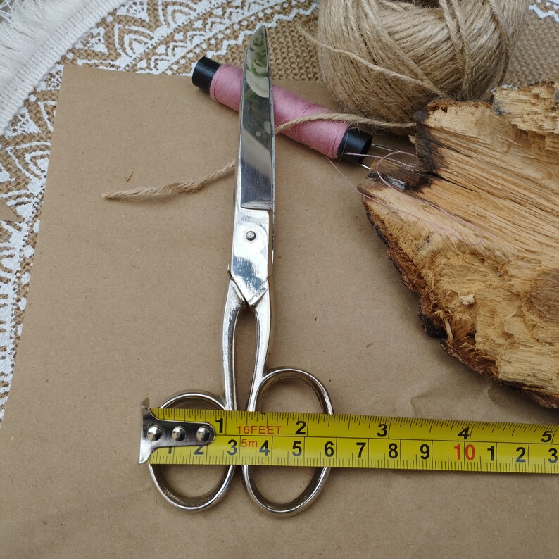 May include: Silver scissors with a yellow measuring tape wrapped around the handles. The tape reads "16FEET 2 3 5m 4 5 6 7 8 9 10 1 2 3".