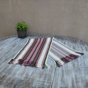 Pode incluir: Caminho de mesa listrado com fundo branco e listras verticais vermelhas e verdes. O caminho de mesa tem bordas com franjas com fios vermelhos, azuis e verdes. &Eacute; exibido em uma mesa de madeira desgastada.
