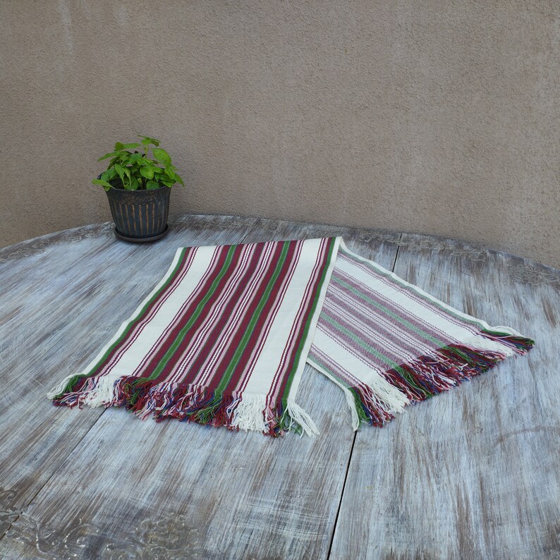 Pode incluir: Caminho de mesa listrado com fundo branco e listras verticais vermelhas e verdes. O caminho de mesa tem bordas com franjas com fios vermelhos, azuis e verdes. &Eacute; exibido em uma mesa de madeira desgastada.