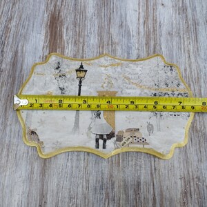May include: A white and gold fabric applique with a winter scene. The applique features a street lamp, a figure in a white dress, and a small building with the text "with love". The applique is approximately 10 inches wide.