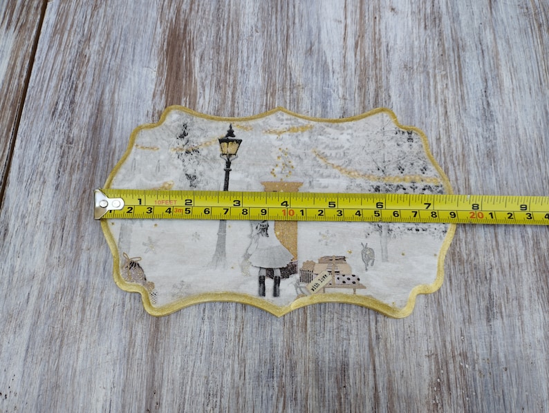 May include: A white and gold fabric applique with a winter scene. The applique features a street lamp, a figure in a white dress, and a small building with the text "with love". The applique is approximately 10 inches wide.
