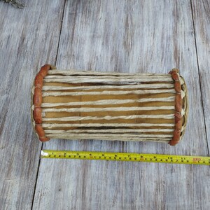 May include: A cylindrical drum with a light brown wooden body and tan leather straps. The drum's ends are bound with brown leather and woven rope. A yellow measuring tape is placed next to the drum, showing a length of approximately 30 cm.