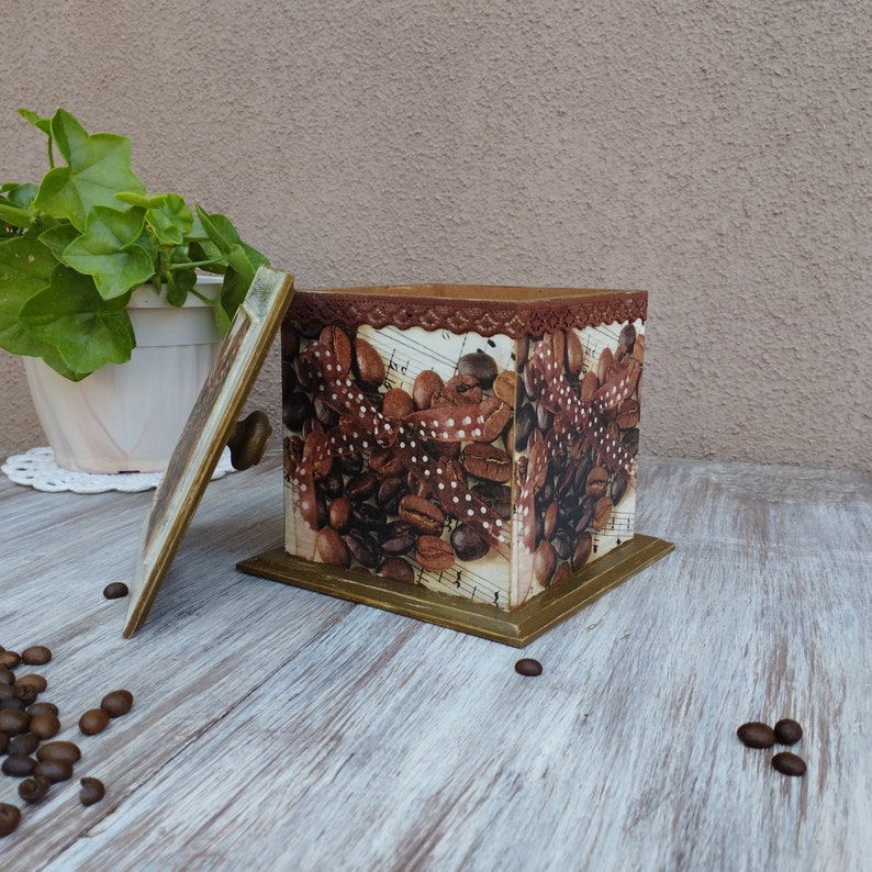 May include: A square wooden box with a hinged lid. The box is decorated with a brown and white pattern of coffee beans and a brown ribbon with white polka dots. The lid has a brown handle. The box is sitting on a light brown wooden surface.