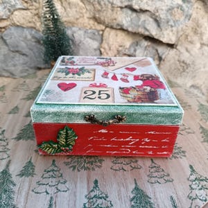 May include: A red and green wooden box with a Christmas theme. The box has a green lid with a white snowy effect and a red base. The lid features a vintage Christmas illustration with a child in a red coat, a basket of gifts, and the number 25. The box has a metal handle and a holly sprig on the side.