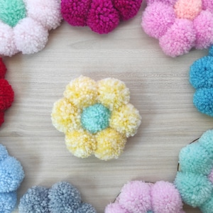 Pode incluir: Uma coleção de flores de pompons de lã coloridas. As flores estão dispostas em círculo sobre uma superfície de madeira. As flores são feitas de lã de diferentes cores, incluindo rosa, azul, amarelo, vermelho e verde.