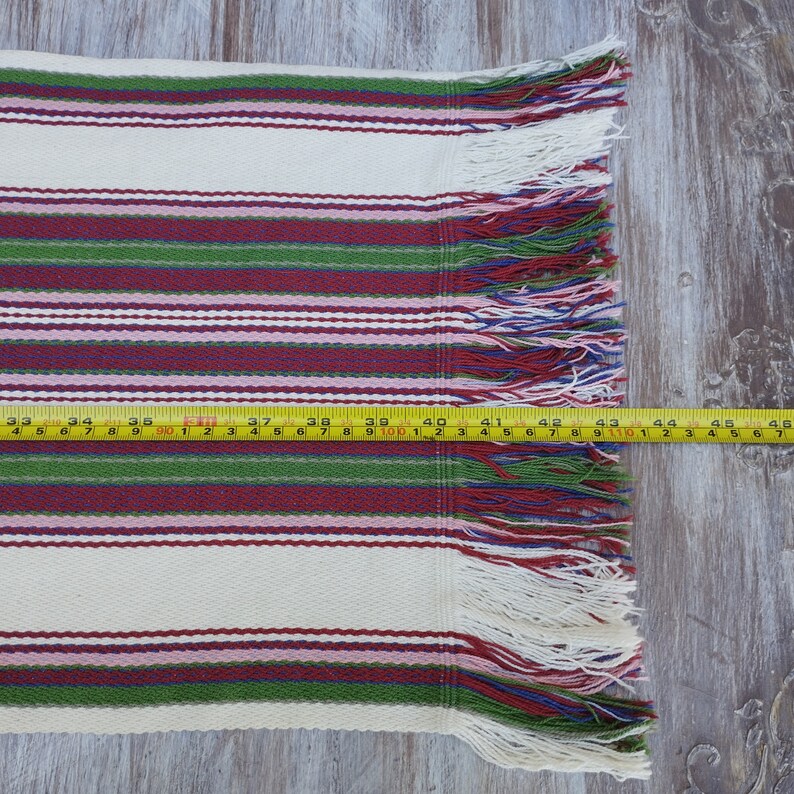 Pode incluir: Um tecido tecido com listras horizontais em creme, bord&ocirc;, verde e rosa. O tecido tem uma borda com franjas e mede aproximadamente 117 cm de comprimento.