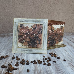 May include: Two wooden boxes with a distressed white paint finish. The boxes are decorated with a collage of coffee beans and musical notes. The boxes are sitting on a light wood surface.