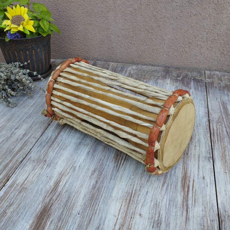 May include: A traditional cylindrical drum with a natural wood finish. The drum's body is wrapped with woven, light-coloured material and secured with leather accents. The drumheads are a light tan colour. This percussion instrument is ideal for music enthusiasts.