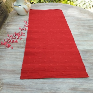 May include: Red table runner with a textured diamond pattern. The runner is made of woven fabric and has a subtle sheen.