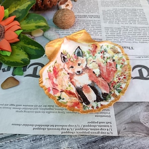 Może przedstawiać: Ręcznie malowana muszla z ilustracją lisa. Lis jest przedstawiony z jesiennymi liśćmi i kwiatową koroną. Muszla ma złoty brzeg, a grafika znajduje się na tle gazety.