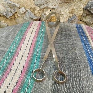 May include: Silver metal tongs with a unique design, featuring two rounded loops at the ends. The tongs are lying on a striped fabric surface.