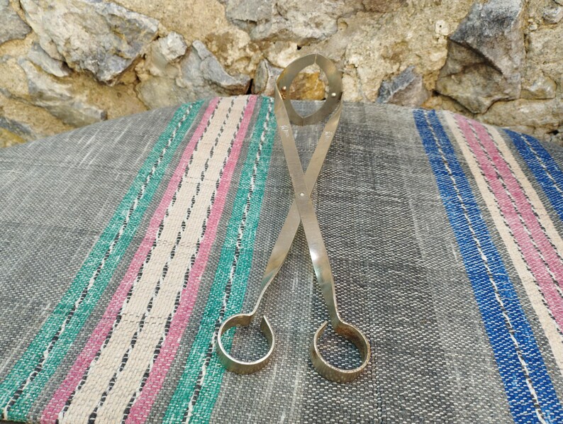May include: Silver metal tongs with a unique design, featuring two rounded loops at the ends. The tongs are lying on a striped fabric surface.
