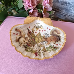 Op de afbeelding: Een witte schelpvormige schaal met een gouden rand, versierd met een geschilderd tafereel van twee eekhoorns in een bossetting met bladeren, eikels en bessen.