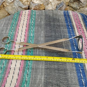 May include: Silver-toned metal tongs with a long handle and two rounded ends. The tongs are lying on a striped fabric surface with a measuring tape next to them.