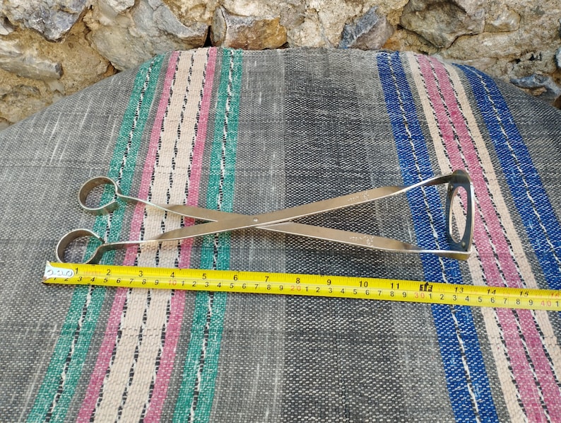 May include: Silver-toned metal tongs with a long handle and two rounded ends. The tongs are lying on a striped fabric surface with a measuring tape next to them.