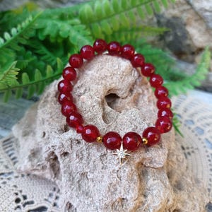 May include: A stretch bracelet composed of faceted deep red beads, interspersed with small metallic gold-toned spacers. A gold-toned star-shaped charm featuring a central clear stone is attached to the bottom of the circular bead strand.