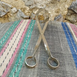 May include: A pair of silver metal tongs with rounded ends. The tongs are crossed and lying on a striped fabric surface.