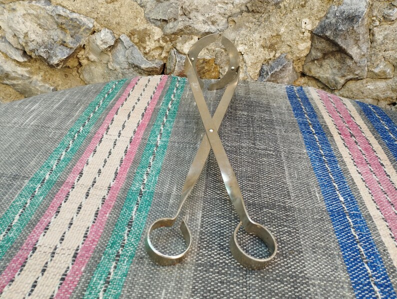 May include: A pair of silver metal tongs with rounded ends. The tongs are crossed and lying on a striped fabric surface.
