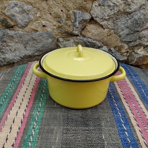 Op de afbeelding: Een gele emaille pan met een zwarte rand en een deksel. De pan heeft twee handvatten en een knop op het deksel.