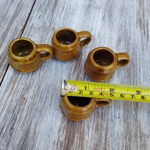 Puede incluir: Cuatro peque&ntilde;as tazas de cer&aacute;mica marr&oacute;n con asas. Las tazas est&aacute;n dispuestas en fila sobre una superficie de madera. Una cinta m&eacute;trica amarilla es parcialmente visible en primer plano.
