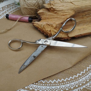 May include: A silver pair of scissors with a wooden background. The scissors have a single blade and a handle with a ring.