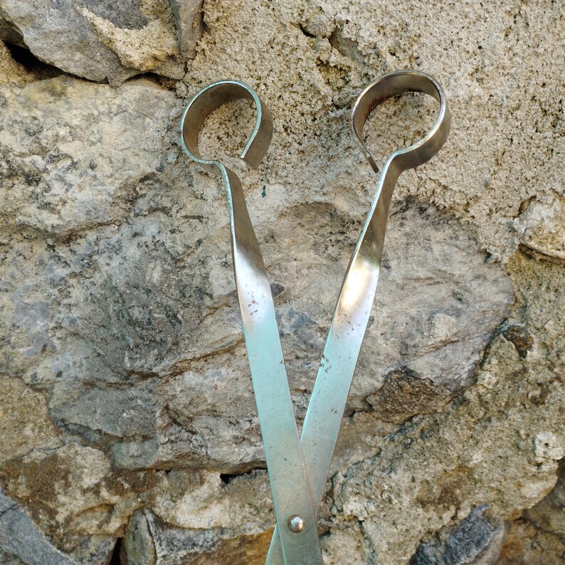 May include: A pair of silver metal tongs with rounded ends. The tongs are open and are resting against a rough, textured stone wall.