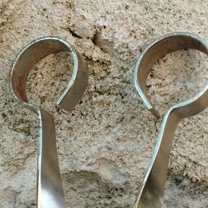 May include: A pair of silver metal tongs with rounded handles. The handles are bent into a U-shape.
